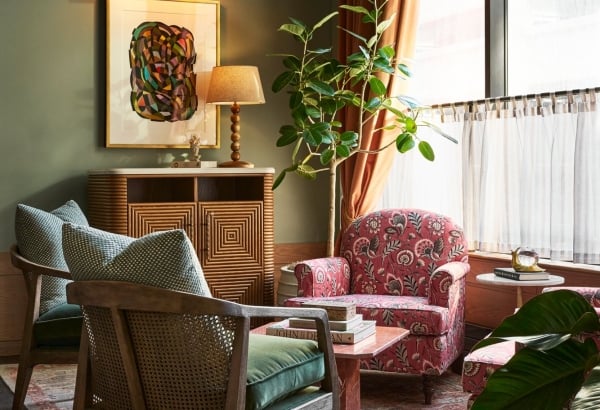 A cozy corner seating area with green walls, large window, and patterned rug features two wooden chairs and two floral armchairs around a pink coffee table. Books: JOHN DEAN, ANNIE LEIBOVITZ.