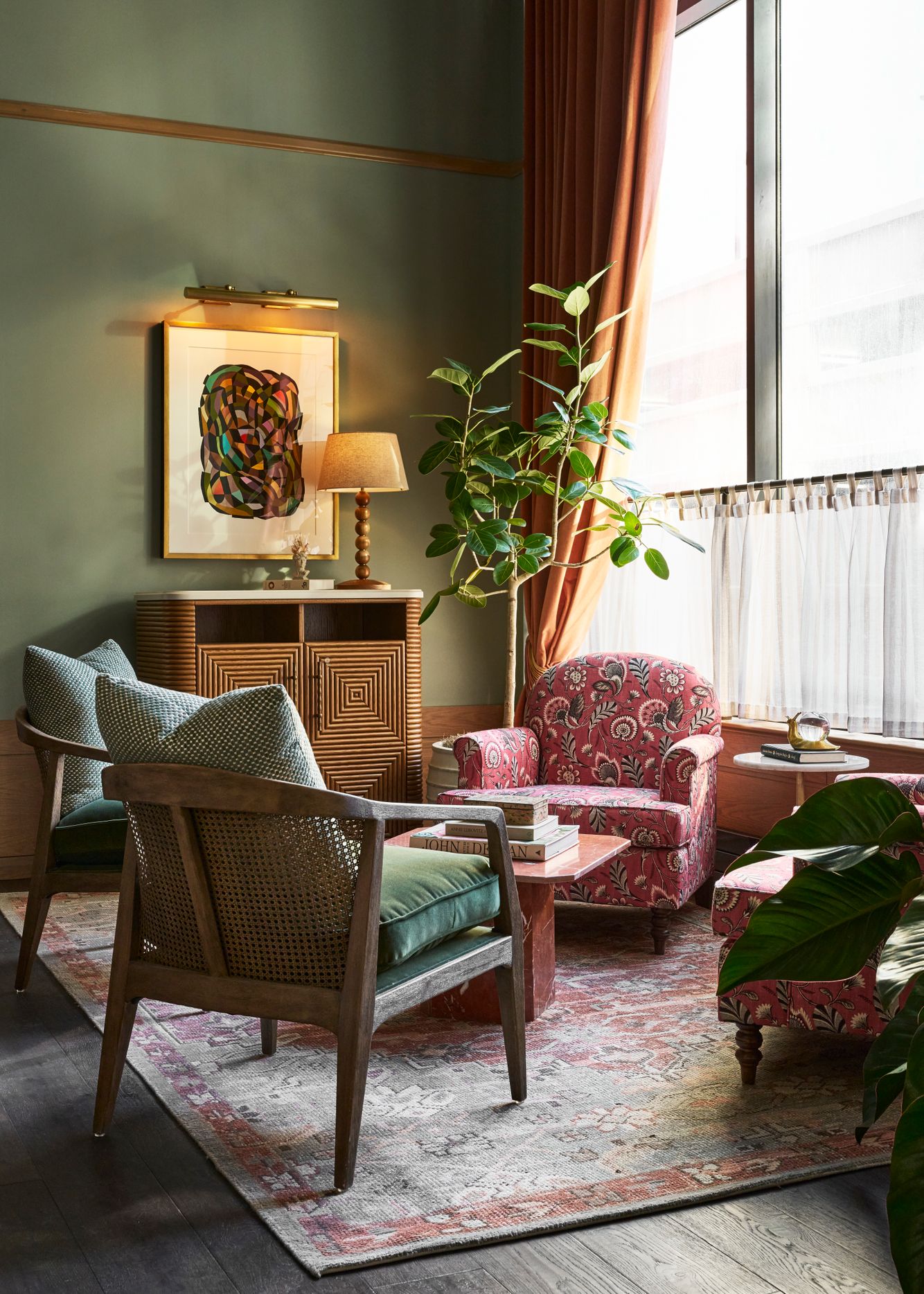 A cozy corner seating area with green walls, large window, and patterned rug features two wooden chairs and two floral armchairs around a pink coffee table. Books: JOHN DEAN, ANNIE LEIBOVITZ.