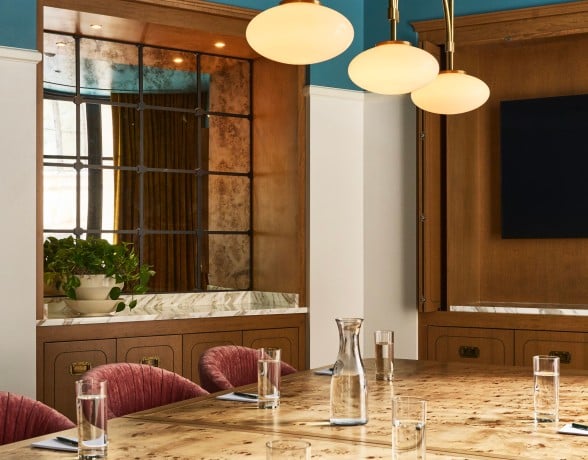 A polished burlwood conference table is set with glasses, carafes, notebooks, and a speakerphone in a room with a blue ceiling and wood-paneled walls.