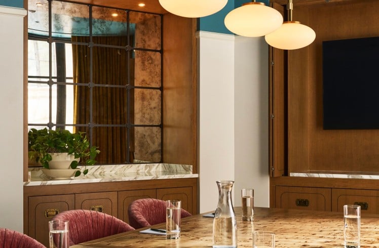 A polished burlwood conference table is set with glasses, carafes, notebooks, and a speakerphone in a room with a blue ceiling and wood-paneled walls.
