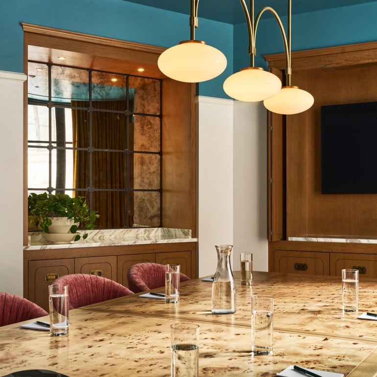 A polished burlwood conference table is set with glasses, carafes, notebooks, and a speakerphone in a room with a blue ceiling and wood-paneled walls.
