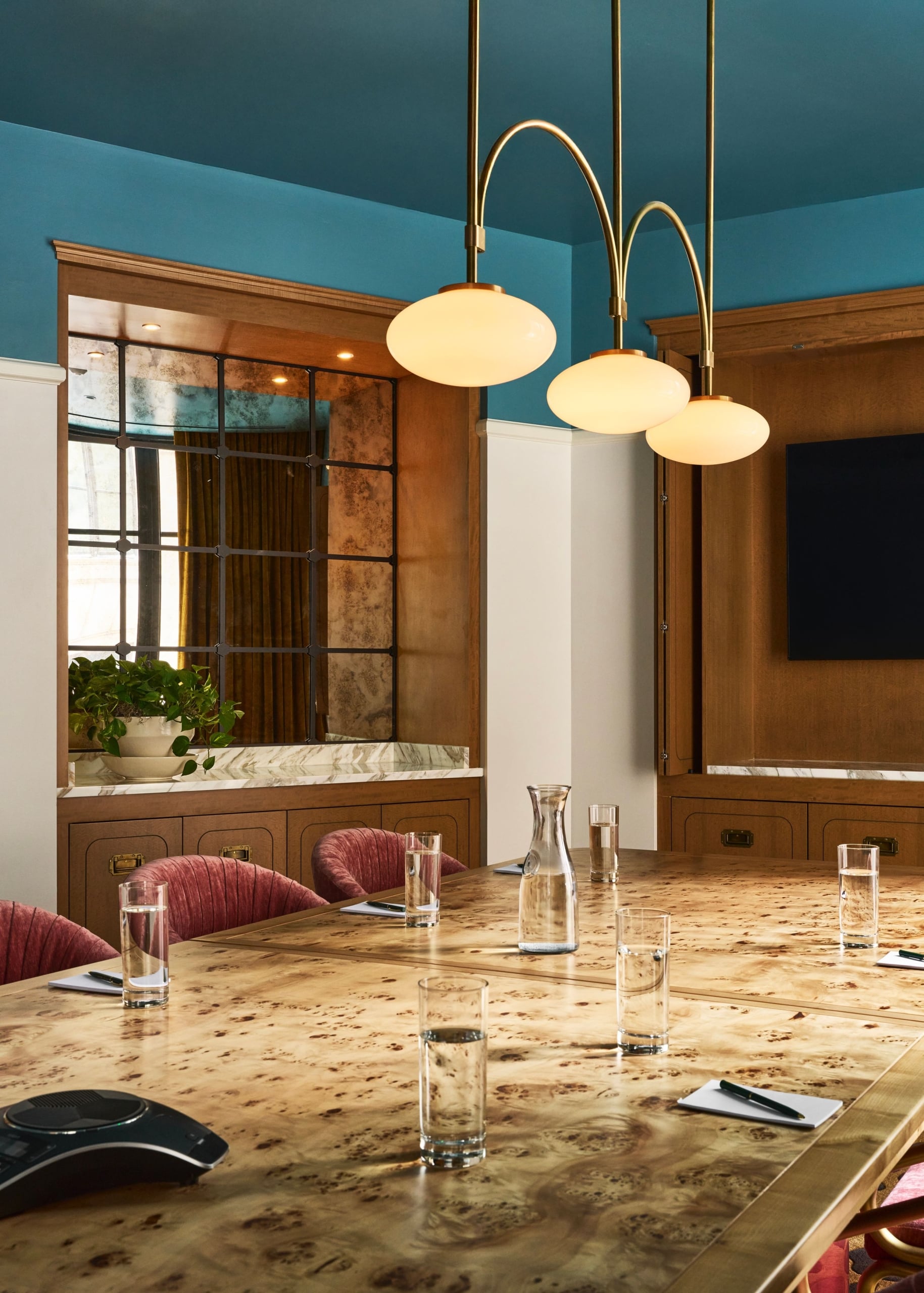 A polished burlwood conference table is set with glasses, carafes, notebooks, and a speakerphone in a room with a blue ceiling and wood-paneled walls.
