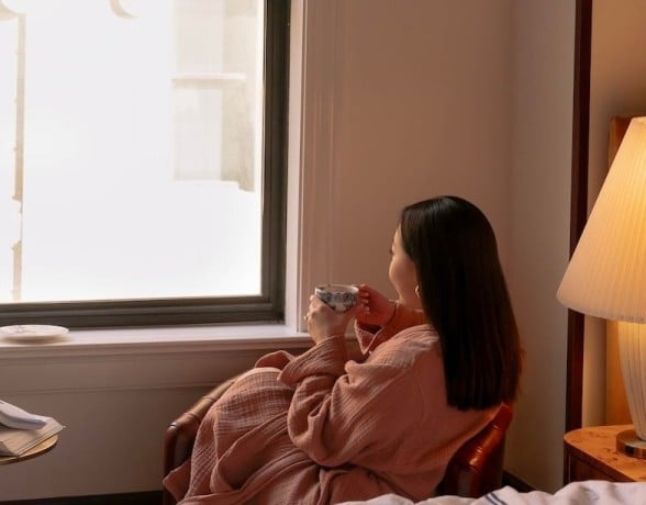 A woman in a peach robe sits, holding a patterned cup, looking out a bright window in a cozy room with a bed and a bedside lamp.