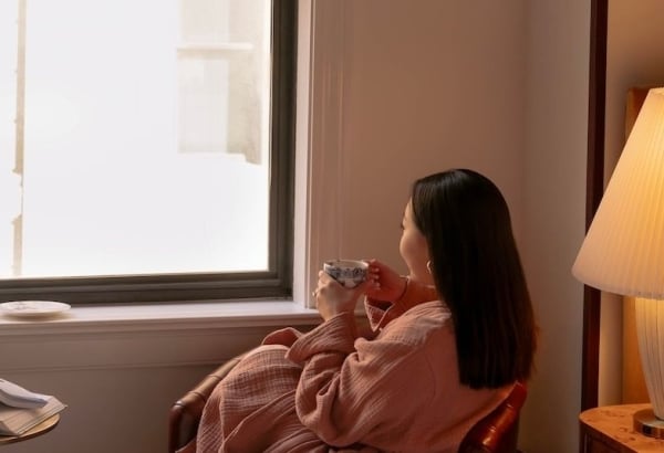 A woman in a peach robe sits, holding a patterned cup, looking out a bright window in a cozy room with a bed and a bedside lamp.