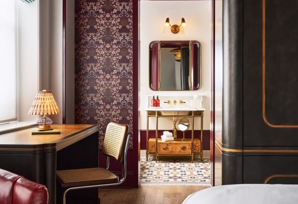 A lavish room with a desk, red armchair, and floral wallpaper. A doorway opens to a bathroom featuring a gold vanity with bottles labeled 'FRESH', 'FACE', and 'HAND', and a patterned tiled floor.