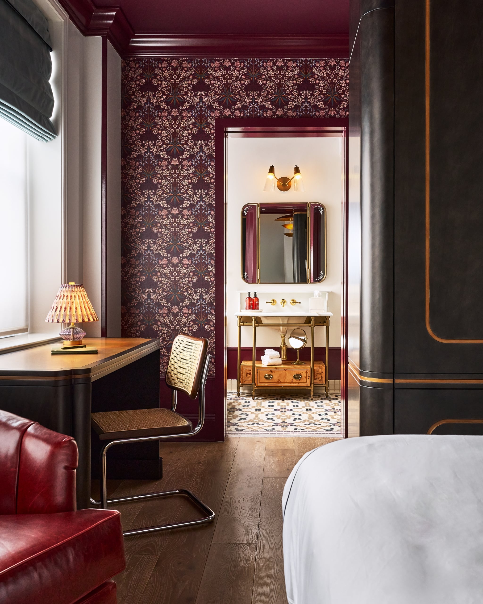 A lavish room with a desk, red armchair, and floral wallpaper. A doorway opens to a bathroom featuring a gold vanity with bottles labeled 'FRESH', 'FACE', and 'HAND', and a patterned tiled floor.