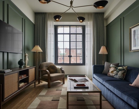 A furnished living room features a blue sofa, armchair, and glass coffee table on a geometric rug. Dark green paneled walls surround a window overlooking city buildings.