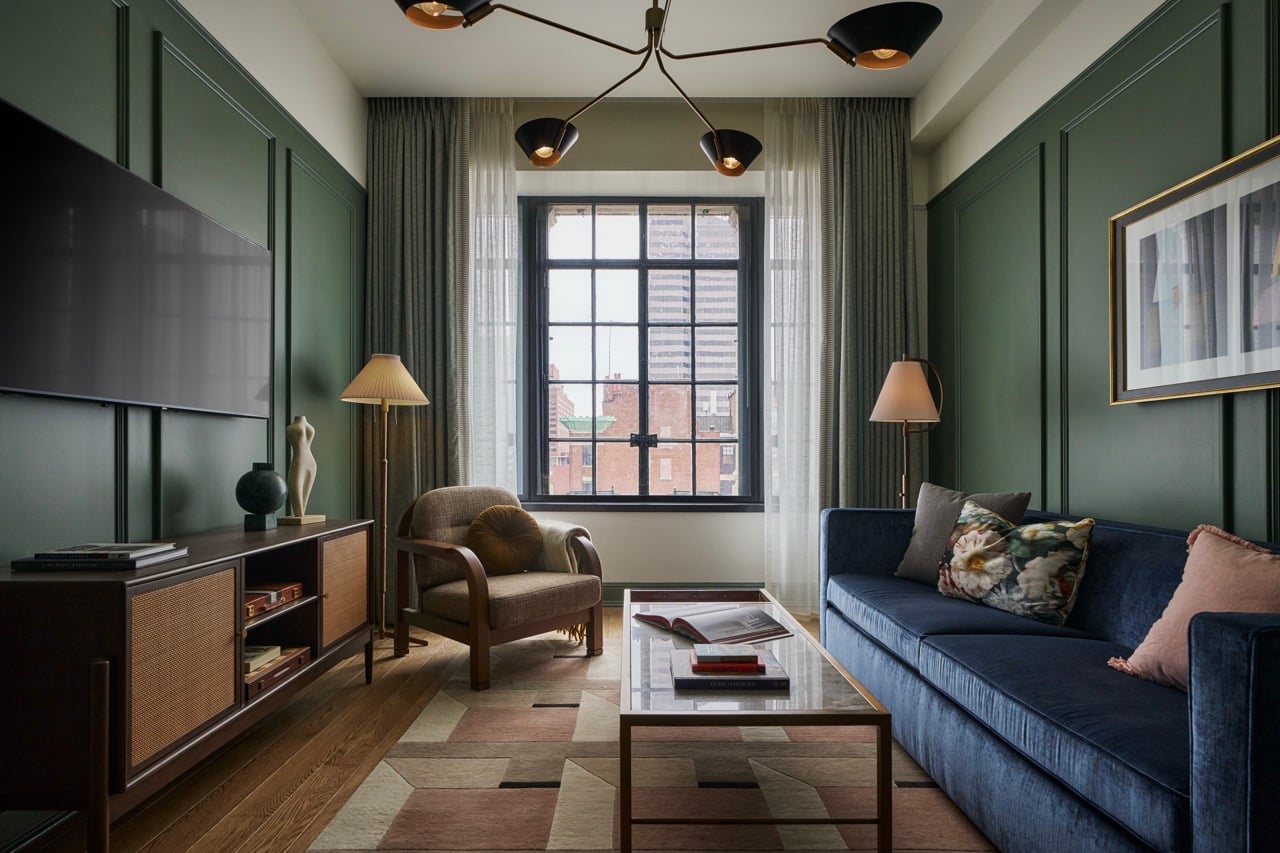 A furnished living room features a blue sofa, armchair, and glass coffee table on a geometric rug. Dark green paneled walls surround a window overlooking city buildings.