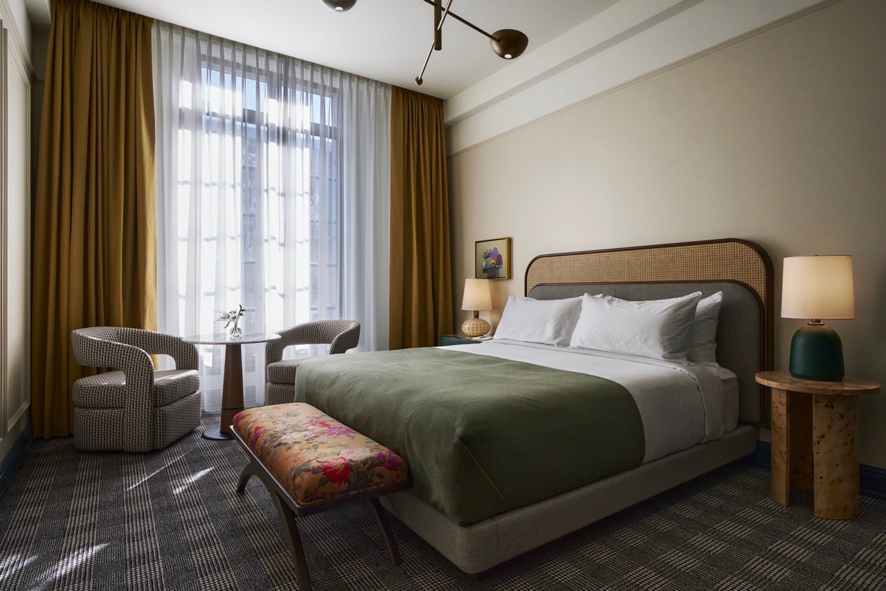 Hotel room features a king bed with rattan headboard, green duvet, and floral bench. A window with gold curtains frames a two-chair table on plaid carpet.