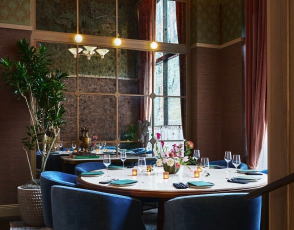 A circular dining table, set with teal napkins and silverware, is surrounded by blue velvet chairs in a luxurious room with patterned walls and a large mirror. Books on a nearby table are titled: "A MAN & HIS CAR", "THE ARCHITECTURE OF TREES".
