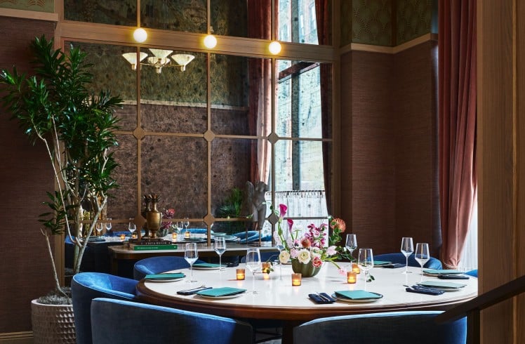 A circular dining table, set with teal napkins and silverware, is surrounded by blue velvet chairs in a luxurious room with patterned walls and a large mirror. Books on a nearby table are titled: "A MAN & HIS CAR", "THE ARCHITECTURE OF TREES".