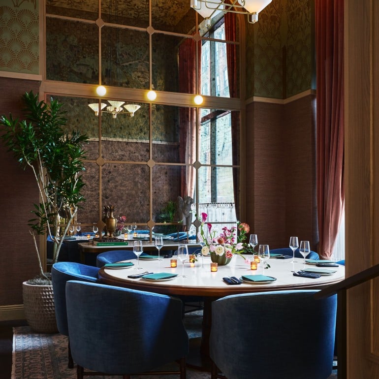 A circular dining table, set with teal napkins and silverware, is surrounded by blue velvet chairs in a luxurious room with patterned walls and a large mirror. Books on a nearby table are titled: "A MAN & HIS CAR", "THE ARCHITECTURE OF TREES".