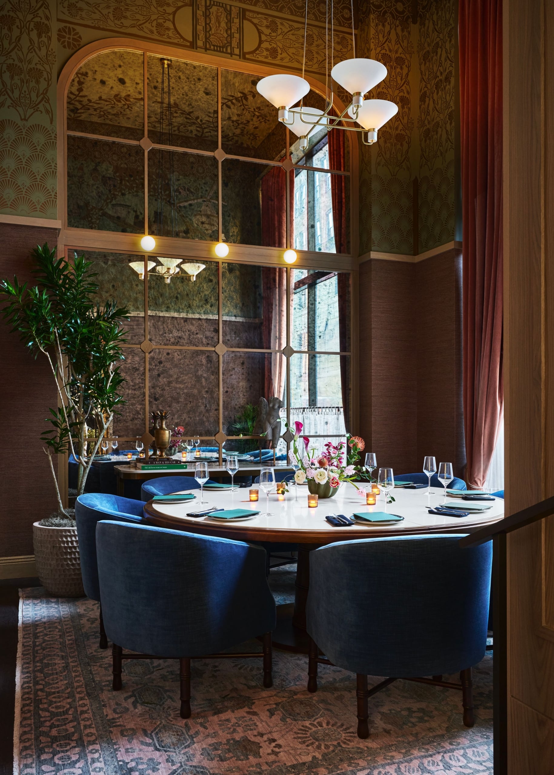 A circular dining table, set with teal napkins and silverware, is surrounded by blue velvet chairs in a luxurious room with patterned walls and a large mirror. Books on a nearby table are titled: "A MAN & HIS CAR", "THE ARCHITECTURE OF TREES".