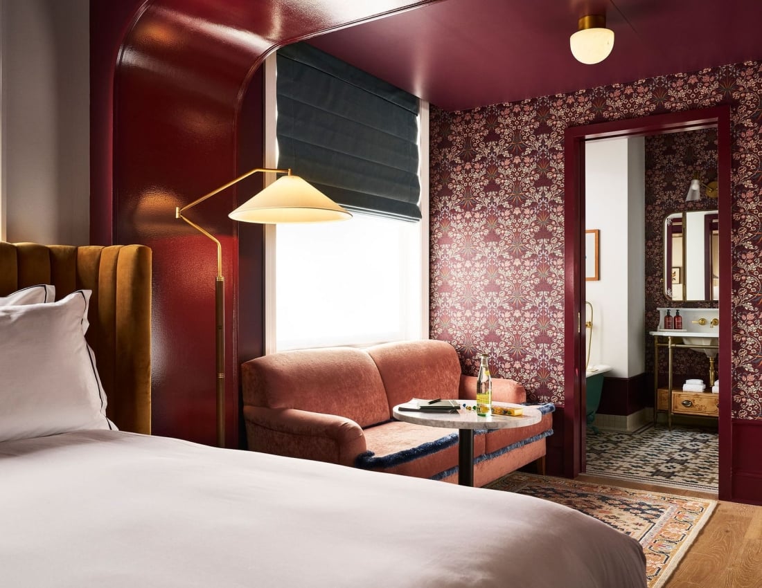 A richly decorated room: A bed with a mustard headboard, a coral armchair by a window with a lamp. Dark floral wallpaper covers walls, leading to a bathroom.