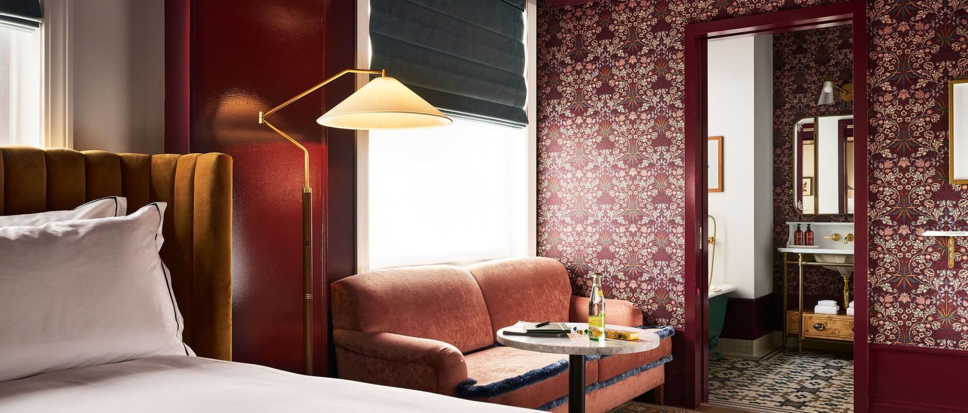 A richly decorated room: A bed with a mustard headboard, a coral armchair by a window with a lamp. Dark floral wallpaper covers walls, leading to a bathroom.