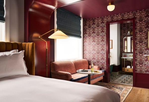 A richly decorated room: A bed with a mustard headboard, a coral armchair by a window with a lamp. Dark floral wallpaper covers walls, leading to a bathroom.