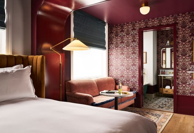 A richly decorated room: A bed with a mustard headboard, a coral armchair by a window with a lamp. Dark floral wallpaper covers walls, leading to a bathroom.