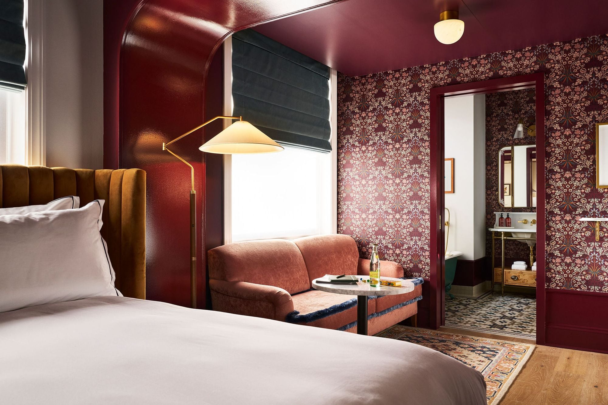 A richly decorated room: A bed with a mustard headboard, a coral armchair by a window with a lamp. Dark floral wallpaper covers walls, leading to a bathroom.