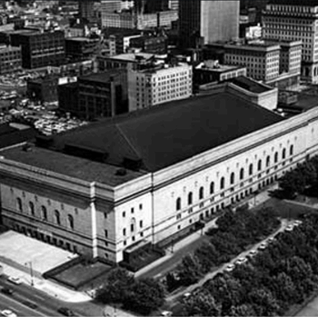 A large, light-colored rectangular building with a dark roof and arched windows dominates a sprawling black-and-white city, flanked by tree-lined streets, cars, and other urban structures.