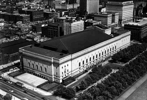 A large, light-colored rectangular building with a dark roof and arched windows dominates a sprawling black-and-white city, flanked by tree-lined streets, cars, and other urban structures.