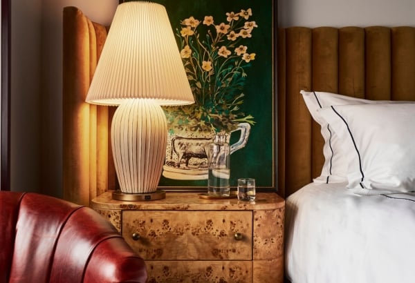 A lit pleated table lamp sits on a burl wood nightstand beside a bed with a velvet headboard and a red leather armchair, beneath a framed floral painting.