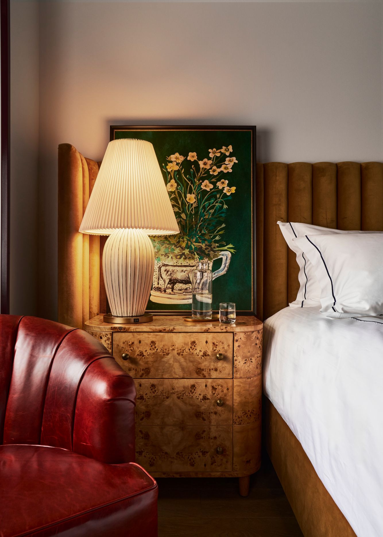 A lit pleated table lamp sits on a burl wood nightstand beside a bed with a velvet headboard and a red leather armchair, beneath a framed floral painting.
