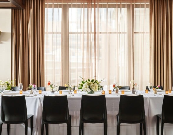 A long dining table is set with place settings, floral arrangements, and votive candles. It is in a room with dark wooden floors and a window wall draped in brown curtains.