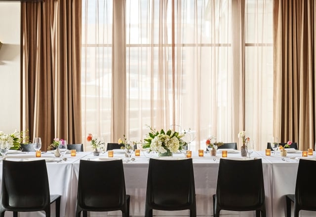 A long dining table is set with place settings, floral arrangements, and votive candles. It is in a room with dark wooden floors and a window wall draped in brown curtains.