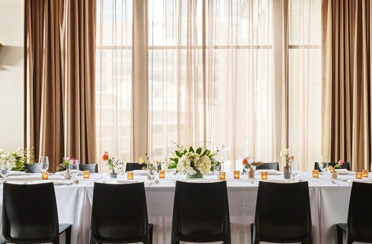 A long dining table is set with place settings, floral arrangements, and votive candles. It is in a room with dark wooden floors and a window wall draped in brown curtains.