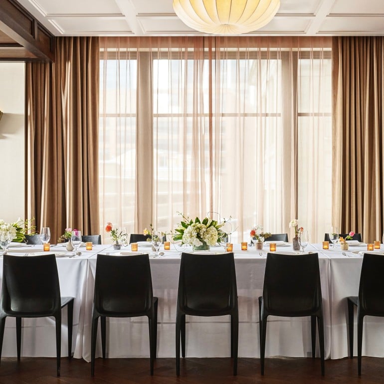 A long dining table is set with place settings, floral arrangements, and votive candles. It is in a room with dark wooden floors and a window wall draped in brown curtains.