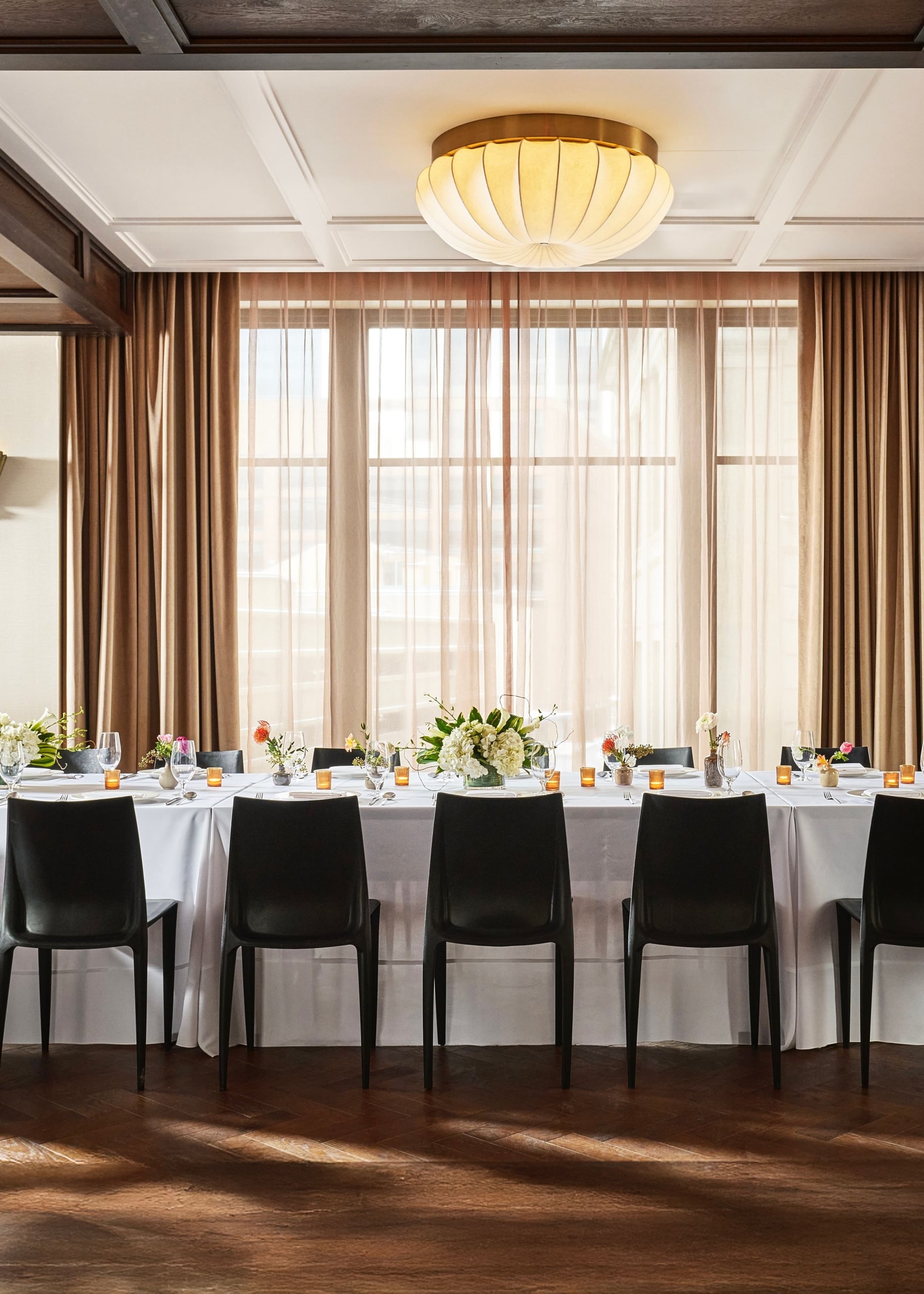 A long dining table is set with place settings, floral arrangements, and votive candles. It is in a room with dark wooden floors and a window wall draped in brown curtains.