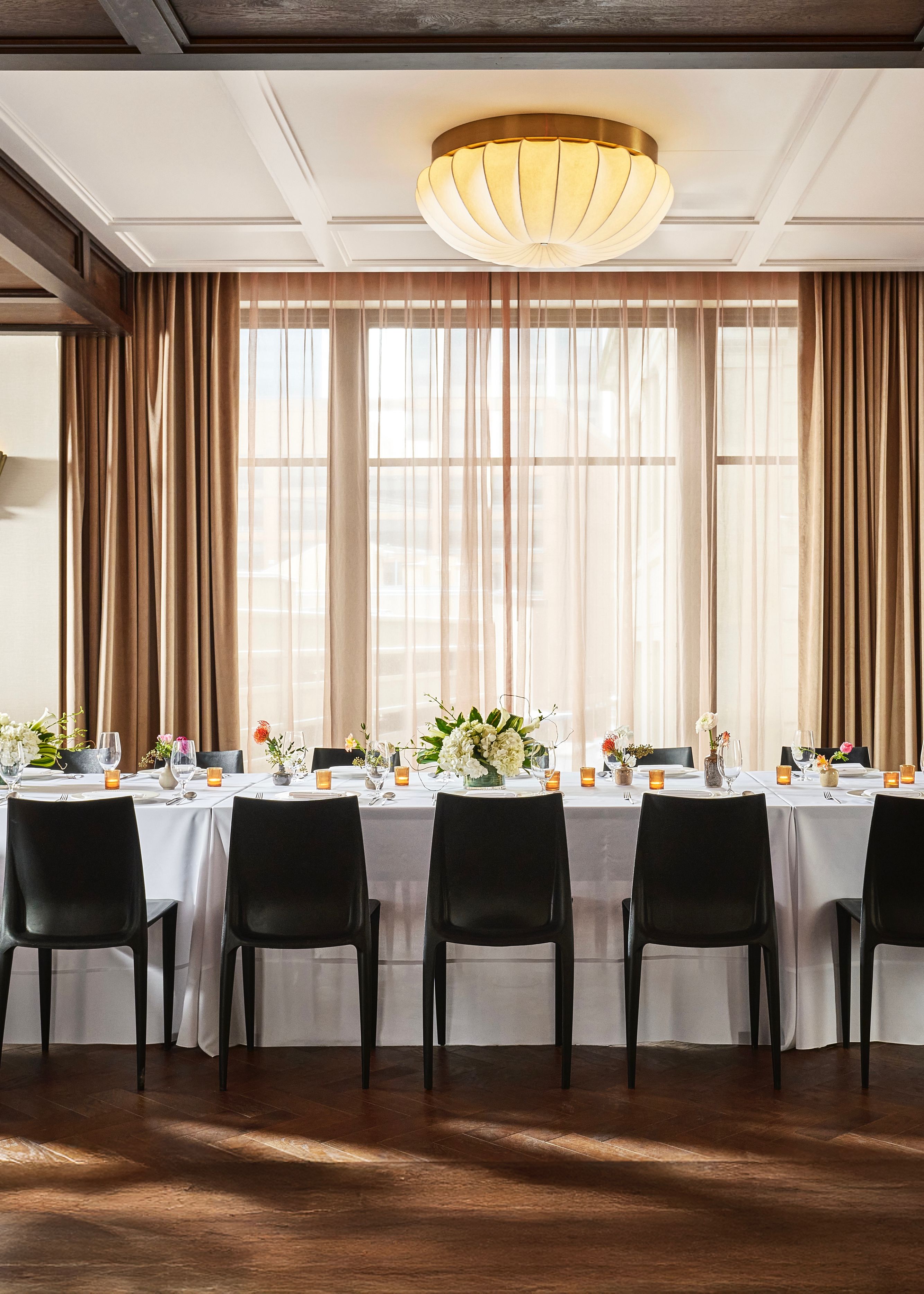A formal dining table set with white linens, flowers, and candles in a room with large windows.