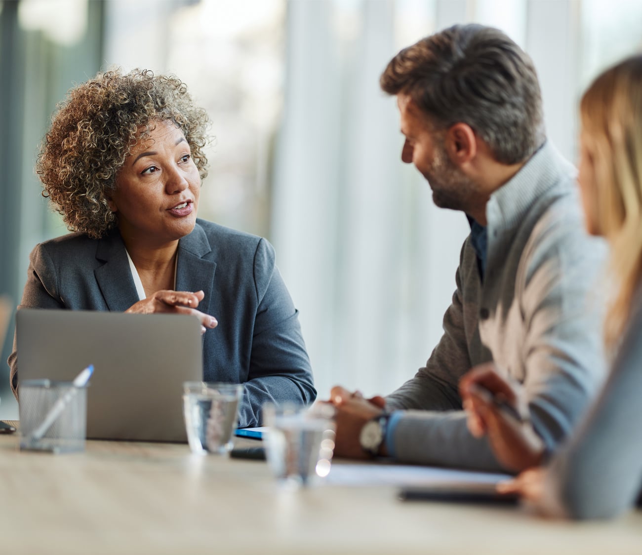 A curly-haired woman in a grey suit speaks, gesturing with hands, at a conference table with a laptop, a man, and a woman present.