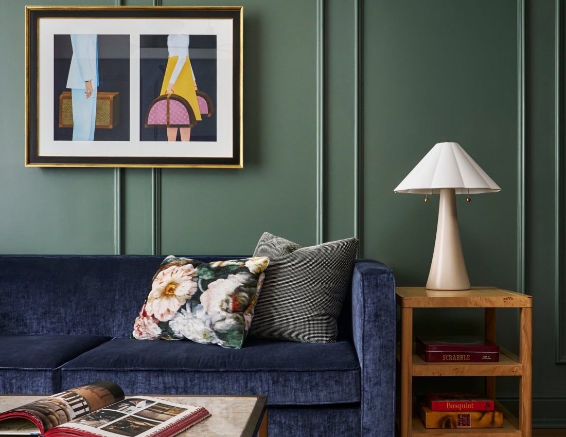 Dark blue velvet sofa with pillows, an open book on a marble coffee table. Dark green paneled wall with framed artwork. Side table holds a lamp and books: SCRABBLE AGES 8+, Basquiat, LOUVRE LA GRANDE COLLECTION.