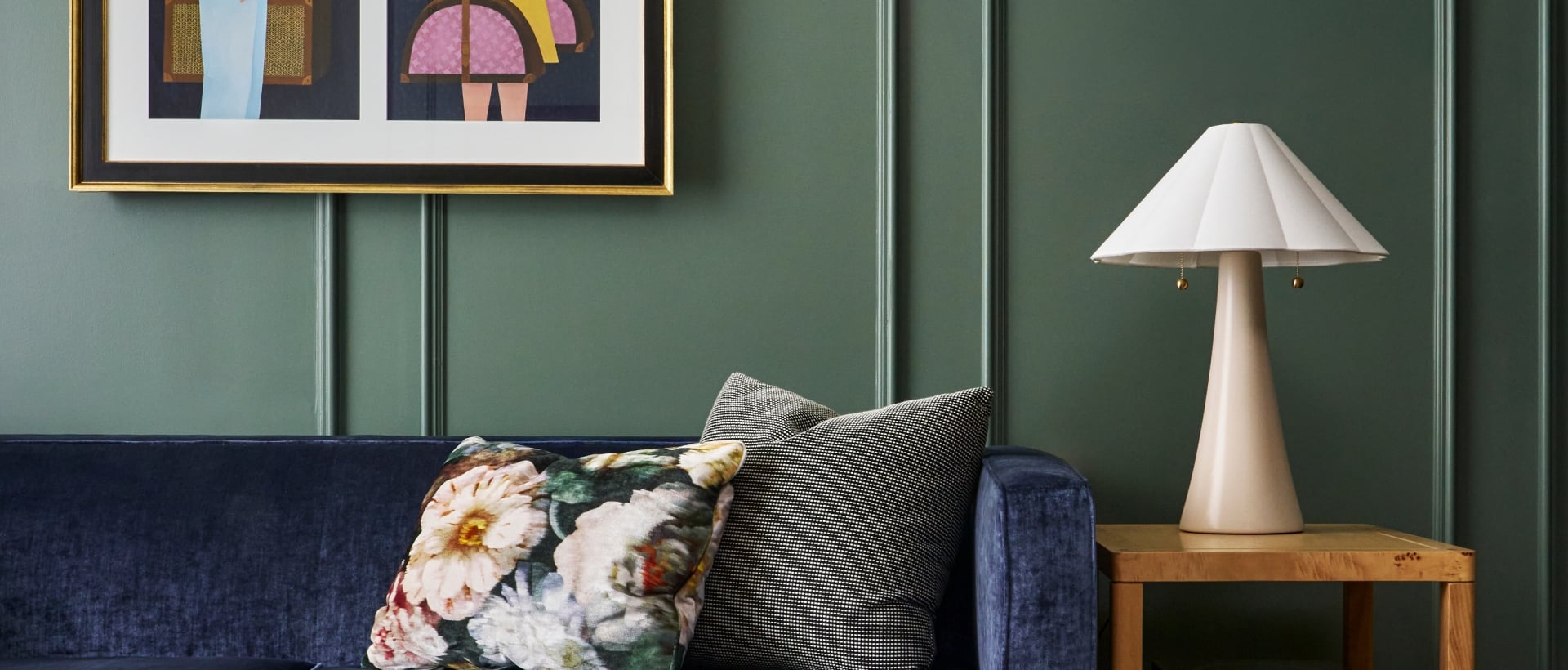 Dark blue velvet sofa with pillows, an open book on a marble coffee table. Dark green paneled wall with framed artwork. Side table holds a lamp and books: SCRABBLE AGES 8+, Basquiat, LOUVRE LA GRANDE COLLECTION.