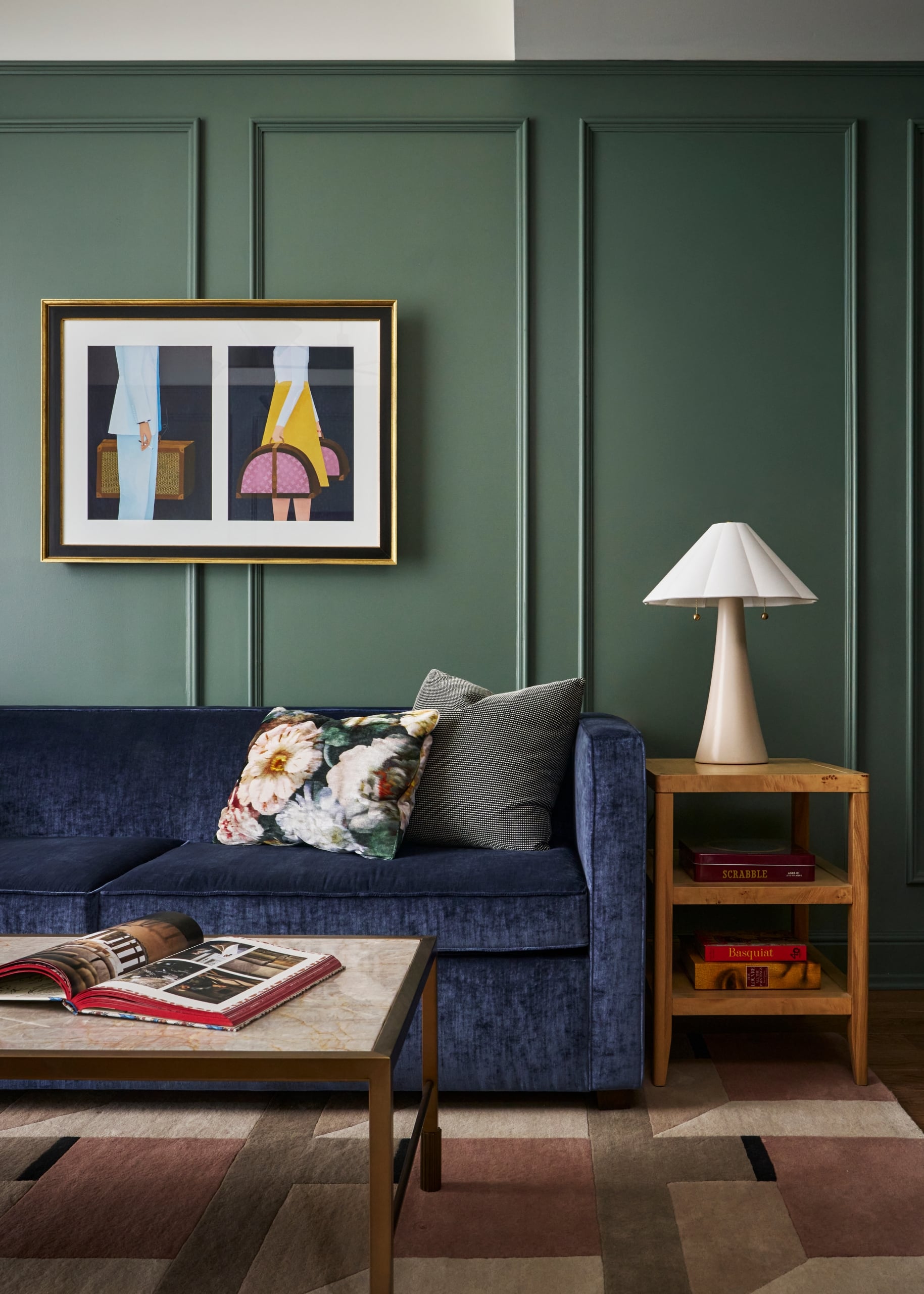 Dark blue velvet sofa with pillows, an open book on a marble coffee table. Dark green paneled wall with framed artwork. Side table holds a lamp and books: SCRABBLE AGES 8+, Basquiat, LOUVRE LA GRANDE COLLECTION.