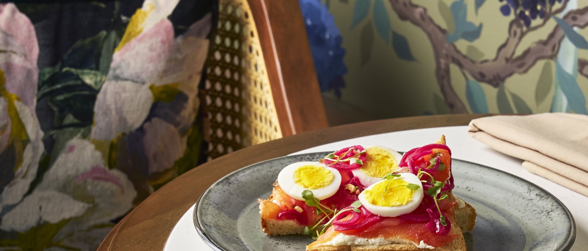 Salmon and egg toasts, coffee, and a fork are on a marble-inlay table. A floral bag rests on a wicker chair against a green wall with purple flowers.