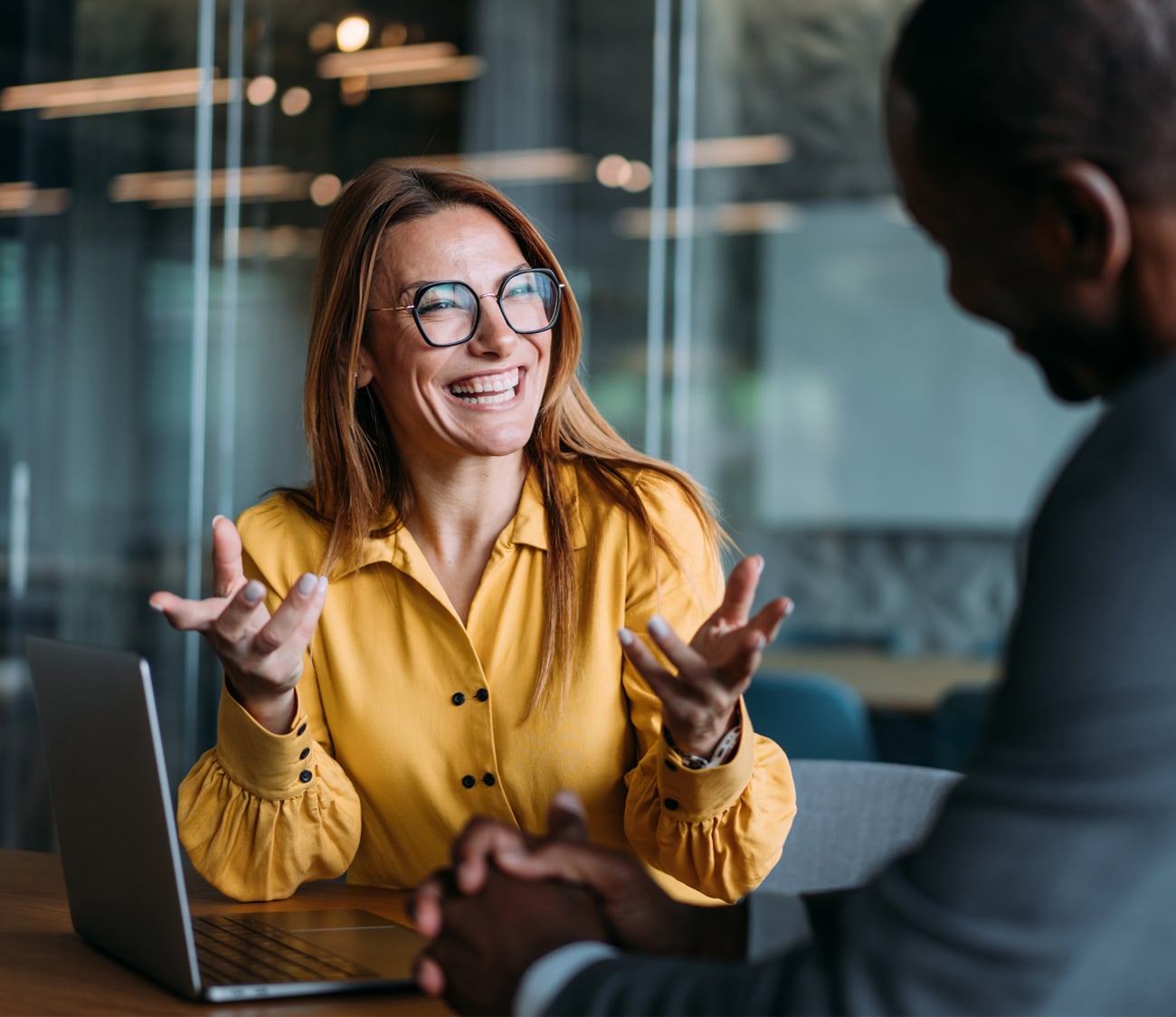A woman in a yellow shirt and glasses laughs, gesturing while talking to a man at a wooden table with a laptop in a modern office setting.