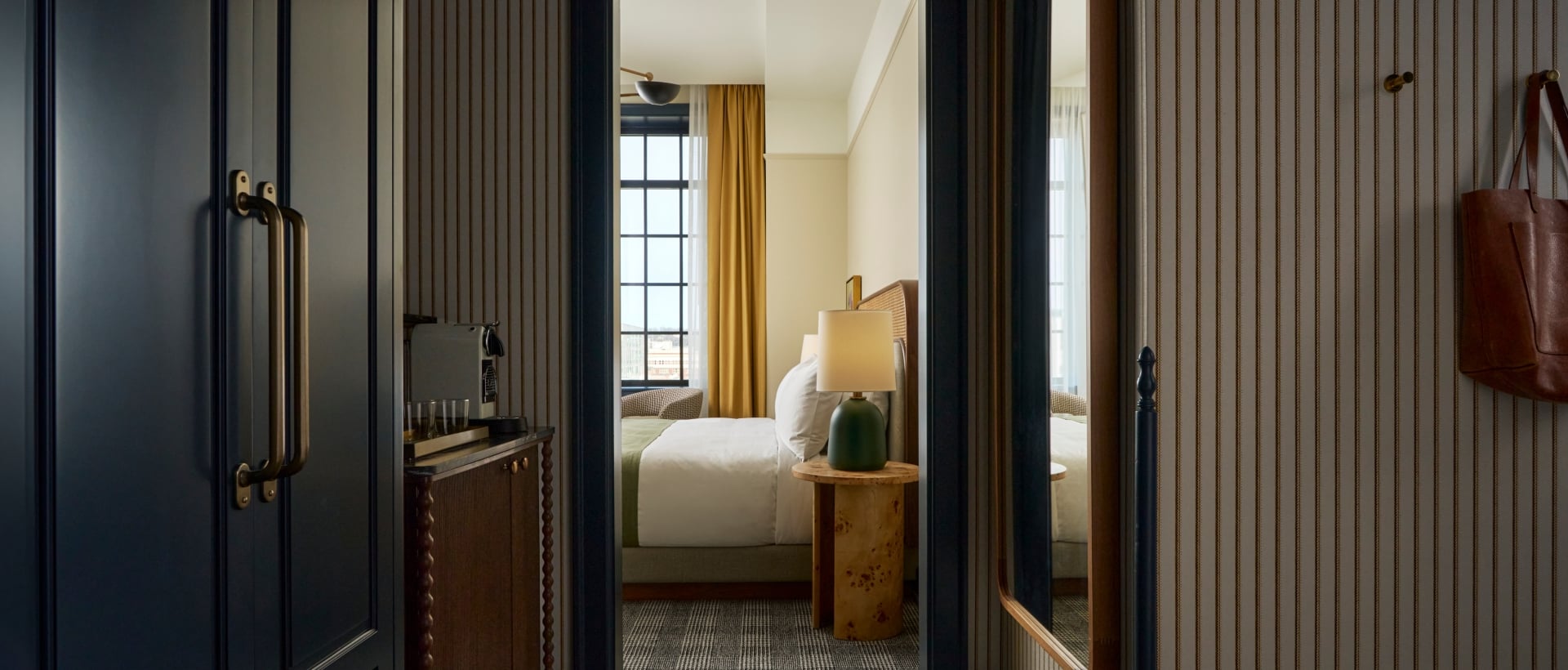 An open doorway reveals a stylish bedroom. A dark hallway with striped wallpaper, plaid carpet, and a coffee station leads to the bright room with a bed and yellow curtains.
