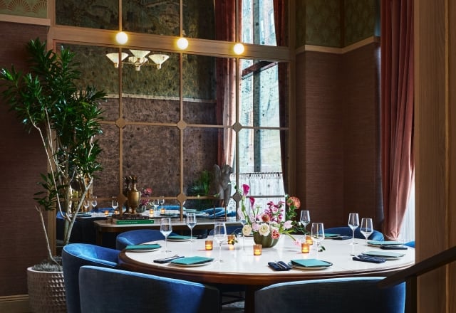 Round dining table formally set with blue place settings, glassware, and a floral centerpiece. Surrounded by blue velvet chairs in an elegant, windowed room with patterned walls. Text found: "A MAN & HIS CAR", "THE ARCHITECTURE OF TREES".