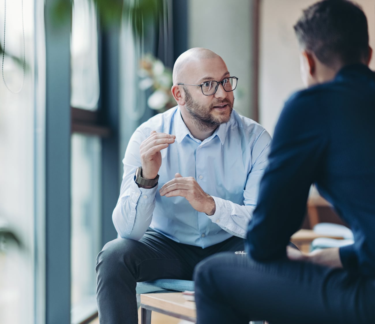 Bald, bearded man in glasses gestures while speaking to another person, seated in a bright office with large windows.