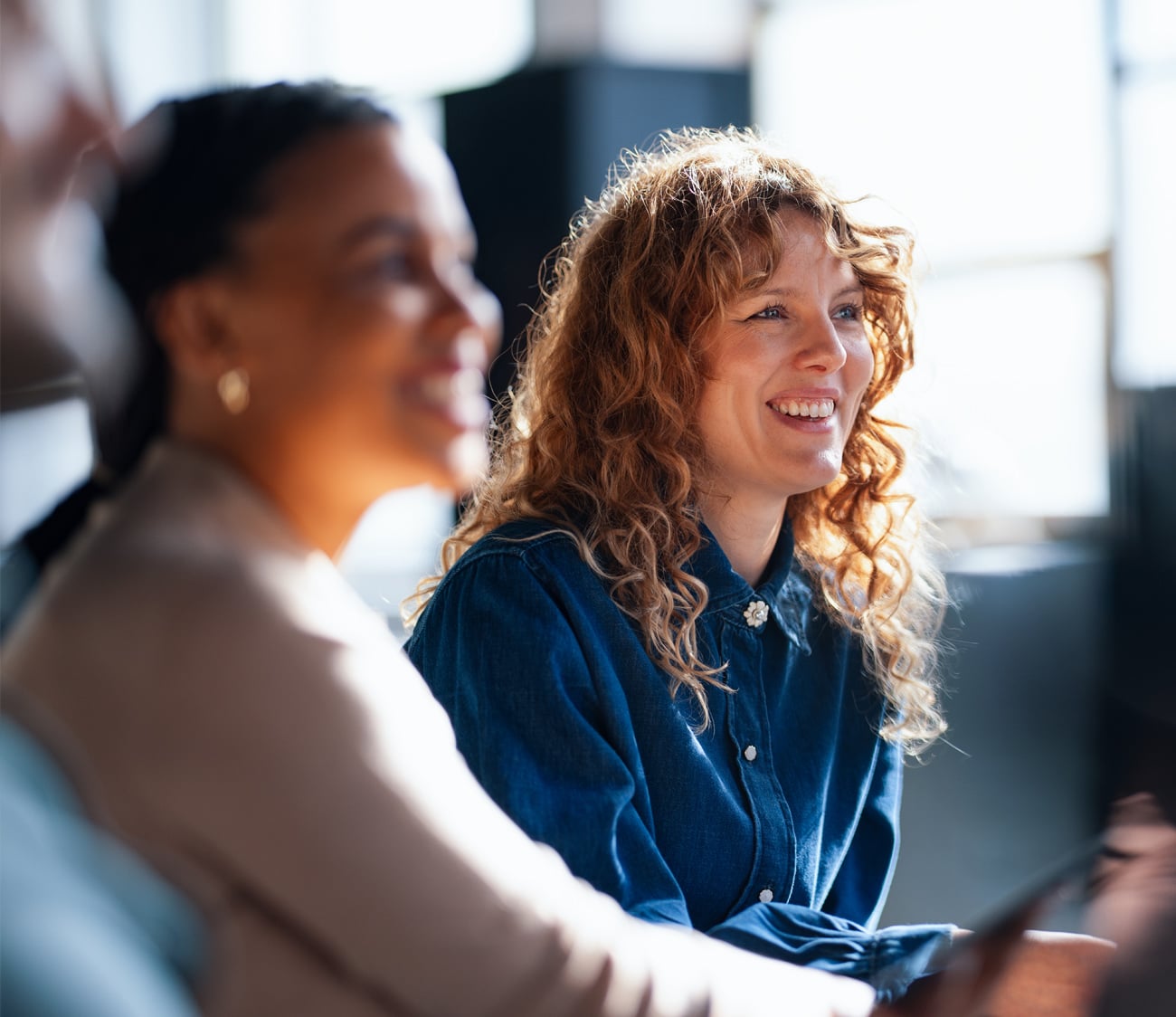 A smiling woman with curly red hair, wearing a denim shirt, looks right, engaged. A blurred person is to her left, with a bright windowed background.