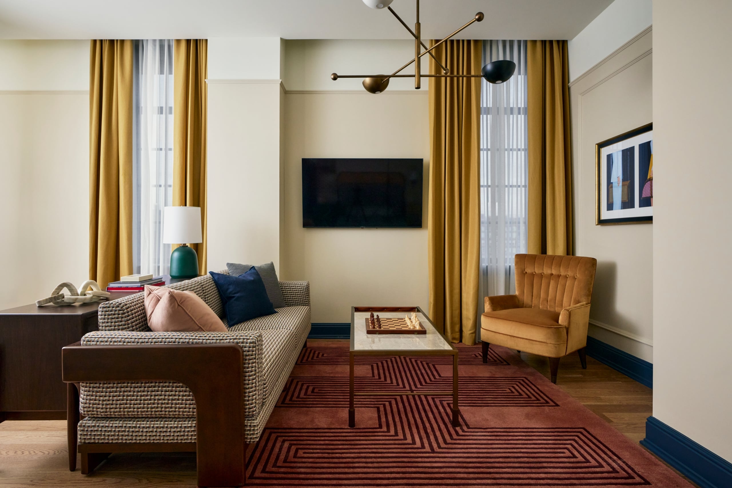 A stylish living room presents a patterned sofa, coffee table with chessboard, and velvet armchair on a geometric rug. Gold curtains frame a window, beside a wall-mounted TV.