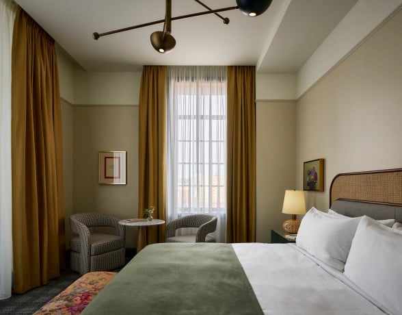 A chic bedroom features a bed with a green throw, two patterned armchairs, and a small table by a window with mustard curtains. Artwork hangs on the walls.