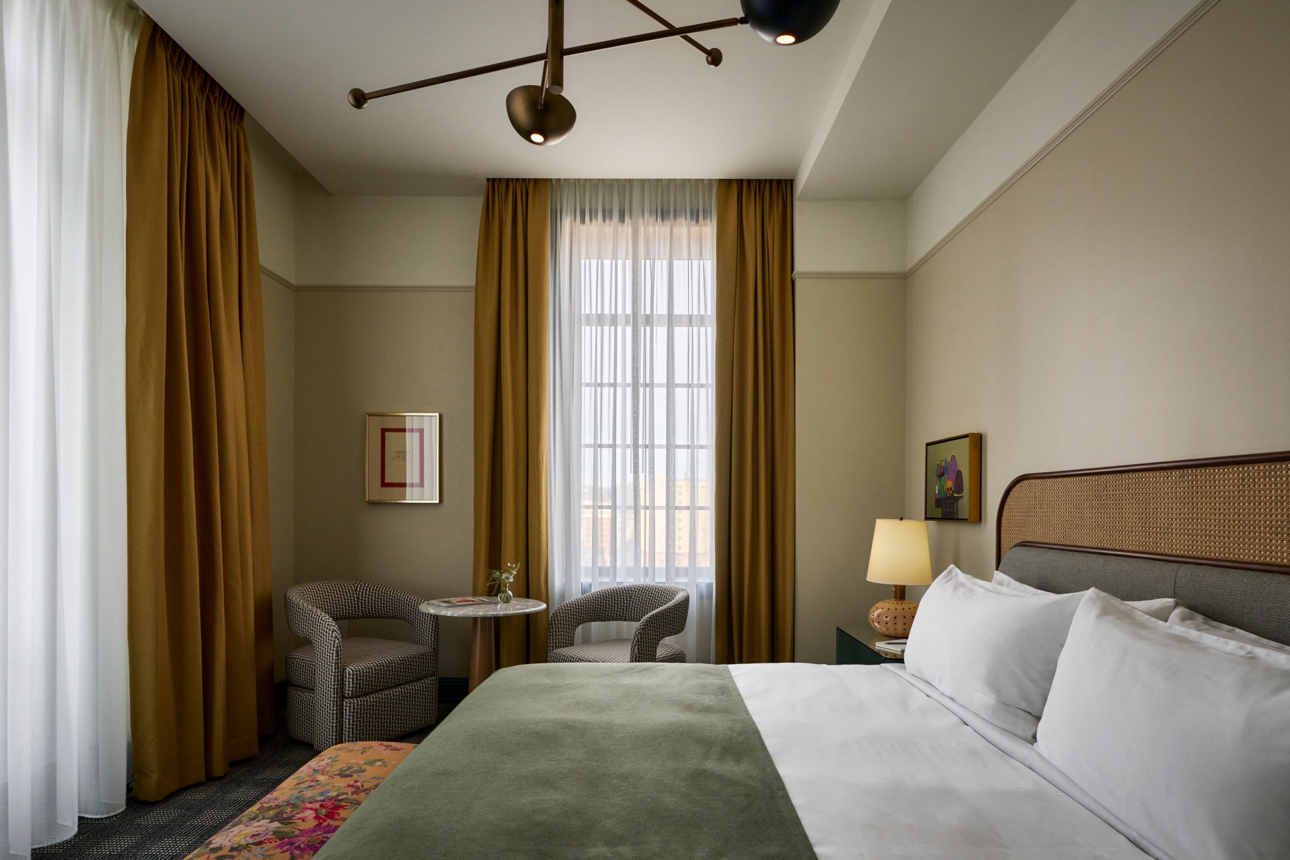 A chic bedroom features a bed with a green throw, two patterned armchairs, and a small table by a window with mustard curtains. Artwork hangs on the walls.