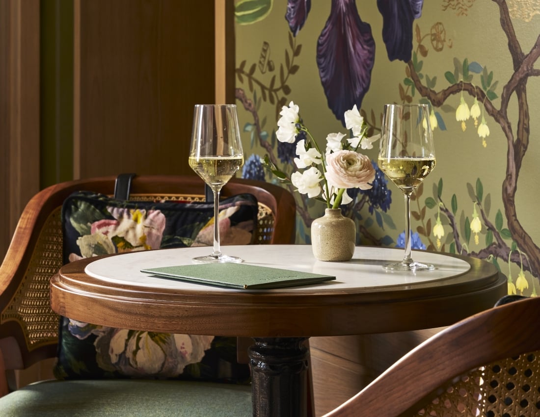 A round table displays two sparkling wine flutes, a vase of flowers, and a green menu reading 'THE LOUNGE'. It sits between two cane chairs against a green wall with botanical wallpaper.