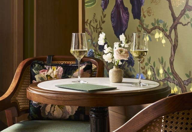 A round table displays two sparkling wine flutes, a vase of flowers, and a green menu reading 'THE LOUNGE'. It sits between two cane chairs against a green wall with botanical wallpaper.