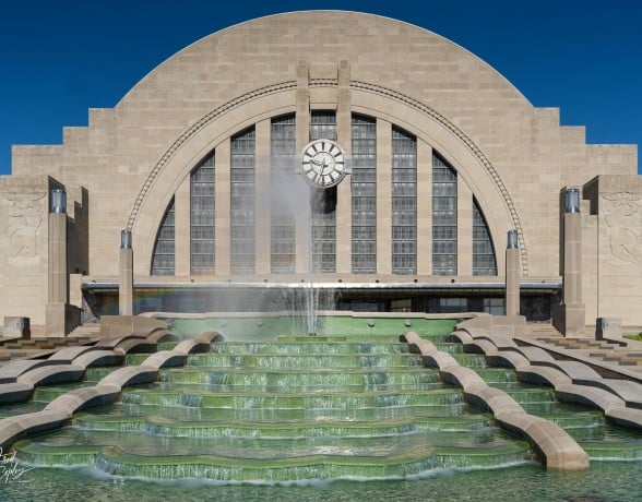 An Art Deco building with a prominent clock and large arched windows overlooks a multi-tiered green fountain with water jets. Fred Copley PHOTOGRAPHY.