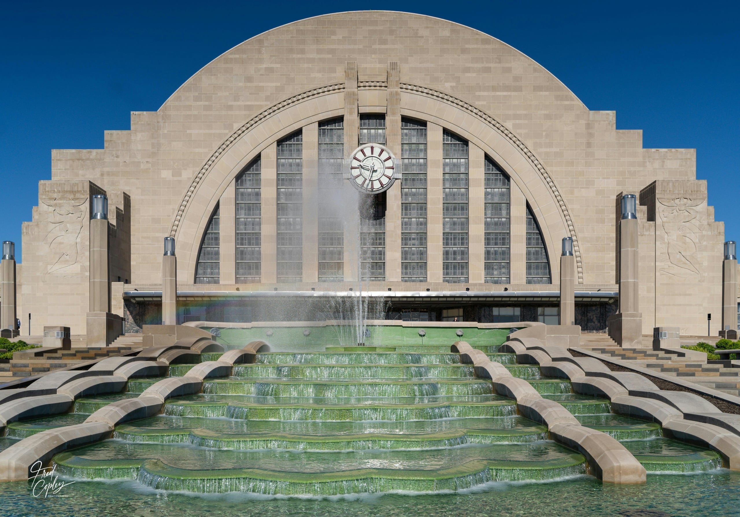 An Art Deco building with a prominent clock and large arched windows overlooks a multi-tiered green fountain with water jets. Fred Copley PHOTOGRAPHY.