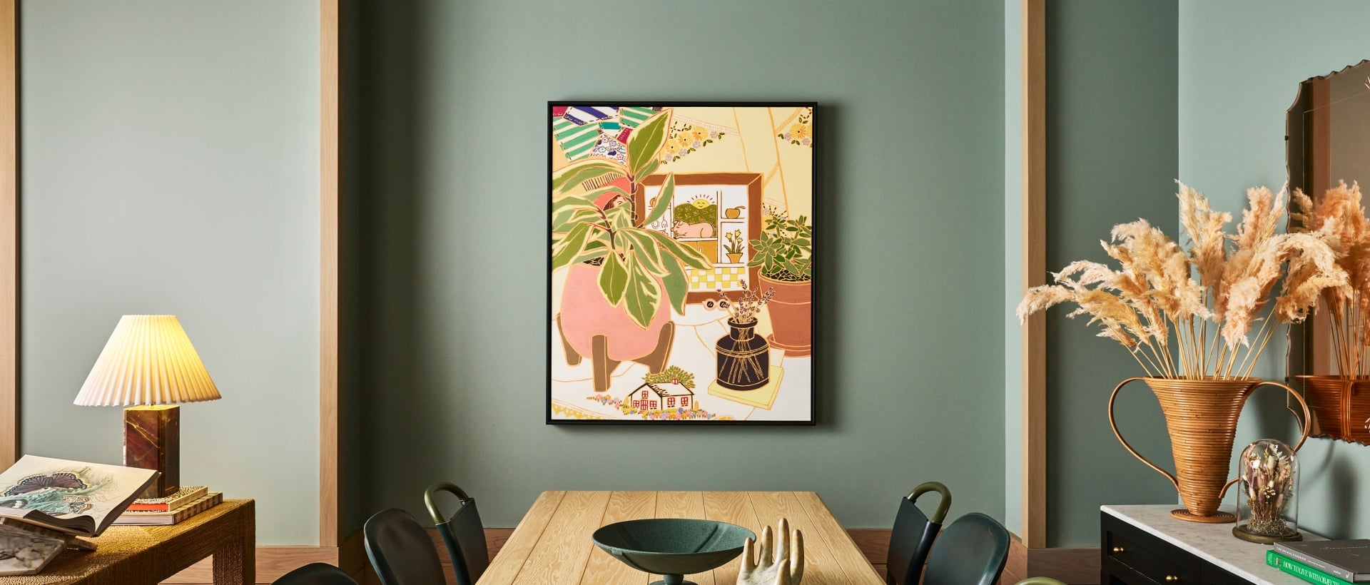 A long wooden dining table with decorative bowls and a hand sculpture sits under a colorful framed artwork on a sage green paneled wall. "HOW TO LIVE WITH OBJECTS" and "THE ANIMAL KINGDOM" books are visible on a side console.